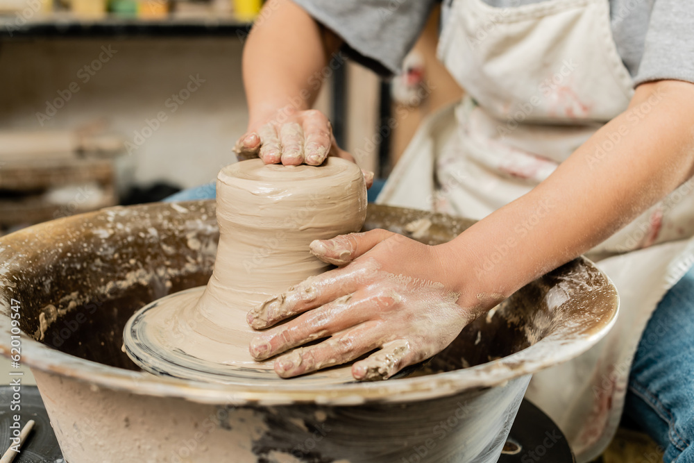 Cropped view of blurred female artisan in apron shaping wet clay and working with pottery wheel in ceramic art workshop at background, skilled pottery making concept Cropped view of blurred female artisan in apron shaping wet clay and working with pottery wheel in ceramic art workshop at background, skilled pottery making concept