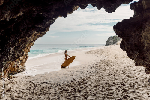 Surf girl with surfboard on ocean beach. Woman with surf board and cave rocks in Bali