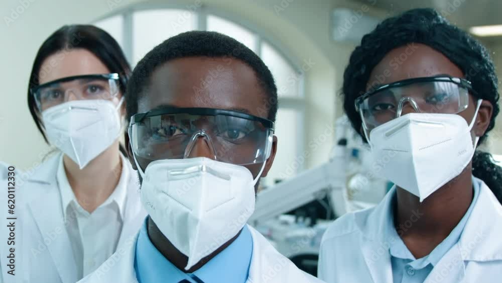 Close up shoot. Three multicultural scientists posing at laboratory room. African American man and woman researchers at foreground. Portrait of lab workers dressed up in special uniform and googles.