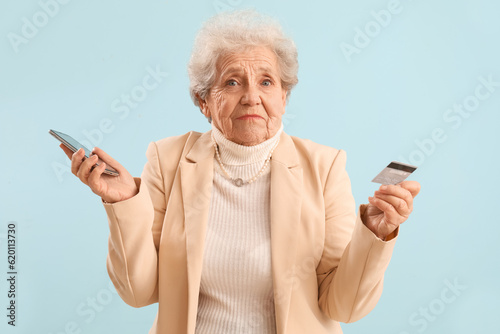 Confused senior woman with mobile phone and credit card on blue background © Pixel-Shot