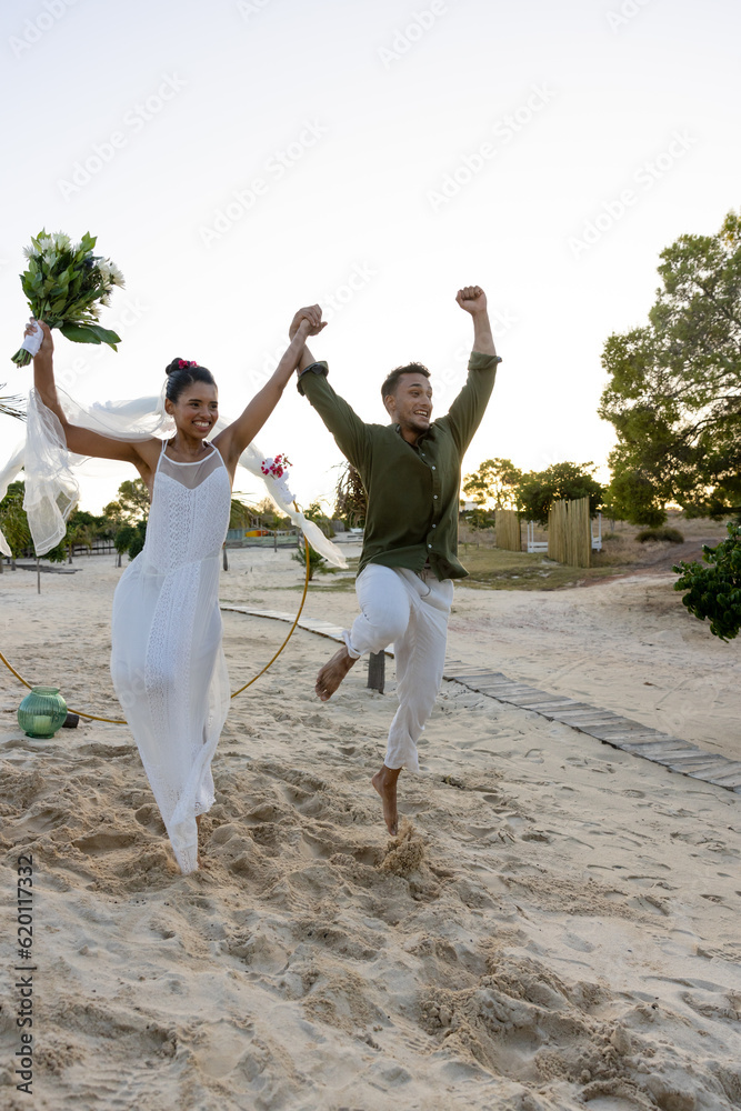 Newlywed biracial couple jumping cheerfully on sandy beach against clear sky at wedding ceremony