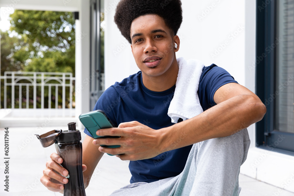 Portrait of happy biracial man using smartphone and earphones sitting on sunny balcony after yoga Portrait of happy biracial man using smartphone and earphones sitting on sunny balcony after yoga