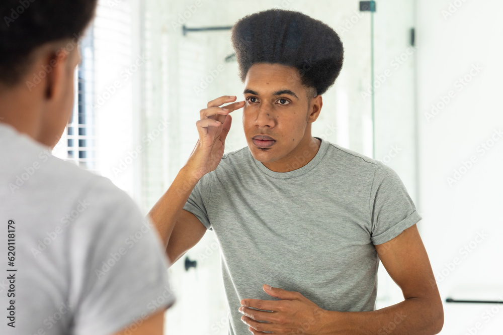 Fototapeta premium Biracial man looking in mirror inspecting face and eyes in sunny bathroom