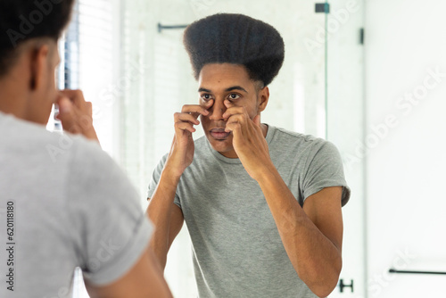 Biracial man looking in mirror inspecting face and eyes in sunny bathroom