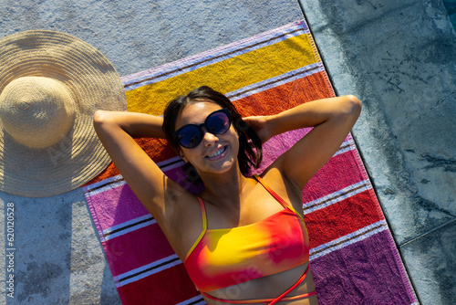 Portrait of happy biracial woman lying on towel and sunbathing next to swimming pool in garden