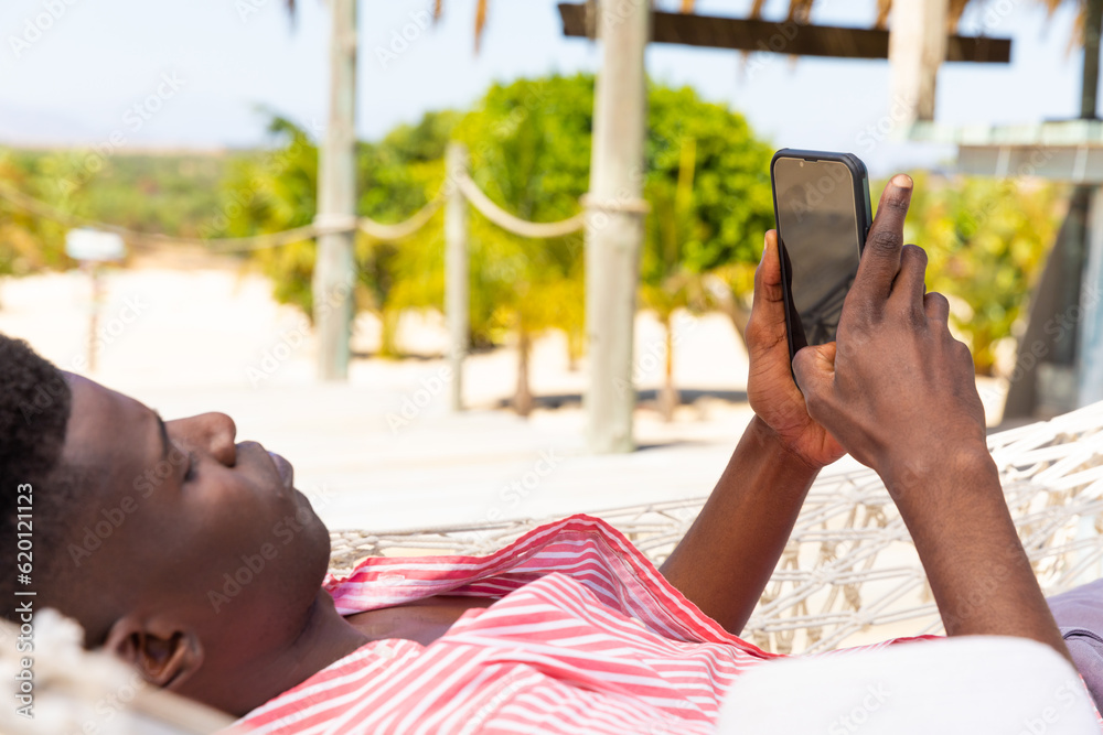 Relaxed biracial man lying in hammock using smartphone at sunny beach bar