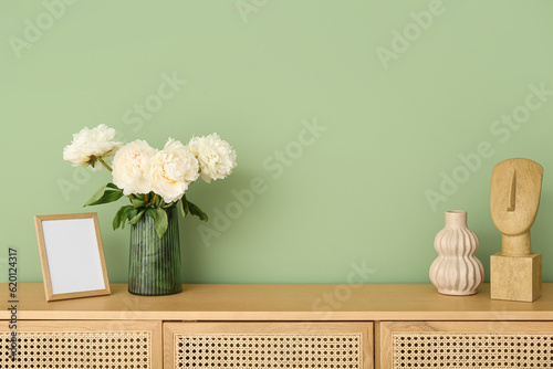 Vase of white peonies with photo frame and figurine on dresser near green wall
