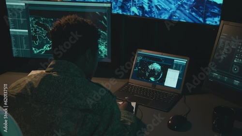 Over-the-shoulder shot of male African American military worker sitting in front of computer screens writing something in notebook