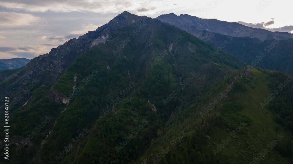 Aerial view of the high valley of the rocky mountain range, on which snow lies in places, near to the fast mountain river at summer cloud morning, blue color of the image