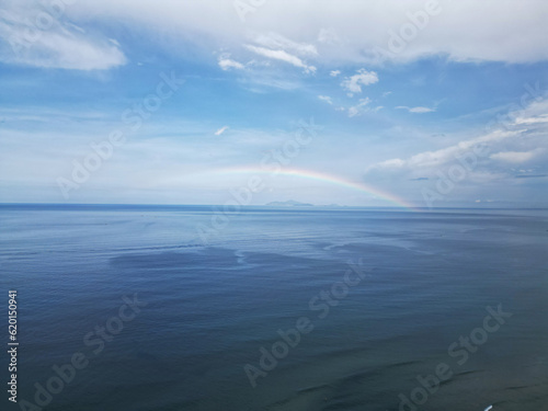 Rainbow around the distant island in the ocean. Like a Disney intro. Cloudy sunny sky after the rain with a deep sea. Small fishing vessels catching fish