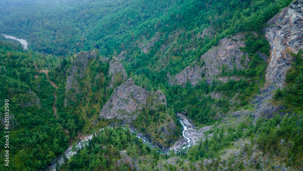 Aerial view of the fast mountain river in valley of the rocky mountain ridges, on which snow lies in places at summer cloud morning, green color of the photo