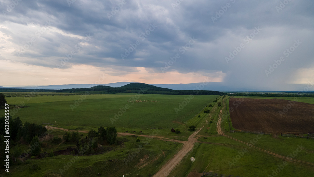 Aerial forward view of the meadows with cows and horses near to the mountain ridge and country road at summer cloud rain day, color