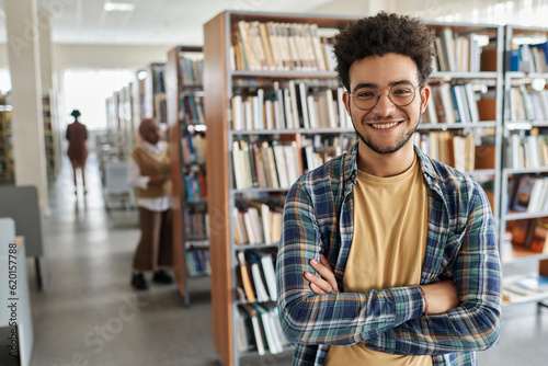 Portrait of student in eyeglasses smiling at camera while standing in library of college