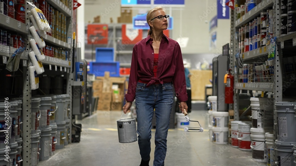 Attractive mature woman carrying can of paint and roller walking down paint aisle in hardware store