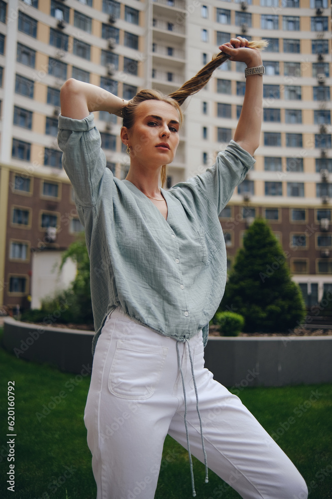 A girl in light summer clothes poses holding her hair against the backdrop of an apartment building.