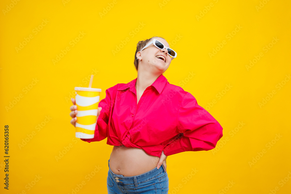 Cheerful pregnant woman in pink shirt holding tasty drink on isolated yellow background. A refreshing drink during pregnancy. A pregnant woman drinks water from a disposable glass Cheerful pregnant woman in pink shirt holding tasty drink on isolated yellow background. A refreshing drink during pregnancy. A pregnant woman drinks water from a disposable glass