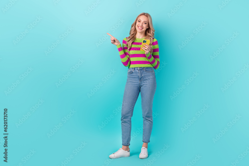 Full size photo of positive woman dressed striped sweatshirt jeans hold smartphone directing empty space isolated on teal color background Full size photo of positive woman dressed striped sweatshirt jeans hold smartphone directing empty space isolated on teal color background