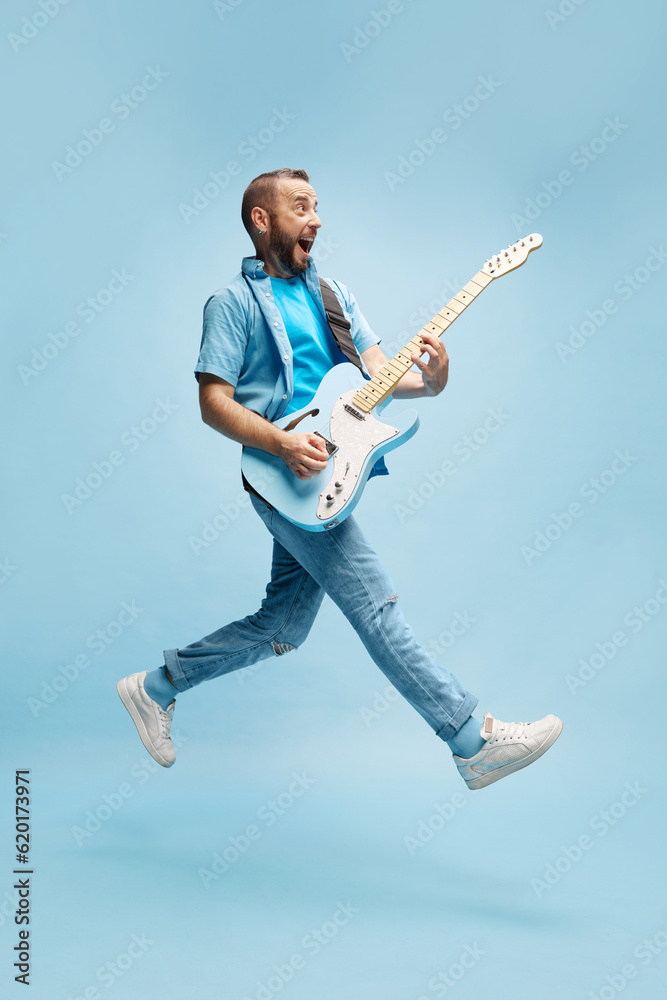 Young, smiling happy man in casual clothes emotionally, cheerfully playing guitar against blue studio background. Concept of music, talent, hobby, entertainment, festival, performance, ad Young, smiling happy man in casual clothes emotionally, cheerfully playing guitar against blue studio background. Concept of music, talent, hobby, entertainment, festival, performance, ad