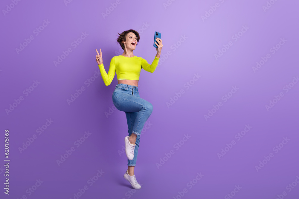Full size photo of excited pretty girl hold smart phone make selfie show v-sign empty space isolated on violet color background