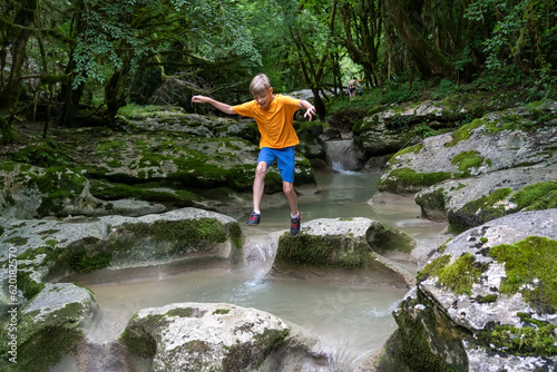 Active boy jumps from rock to rock on a calm mountain river hidden in a dense forest