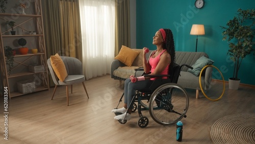 African American woman in a wheelchair exercising with dumbbells in the living room. A woman in a pink headband and in a tracksuit trains the muscles of her arms, shakes her biceps and triceps.