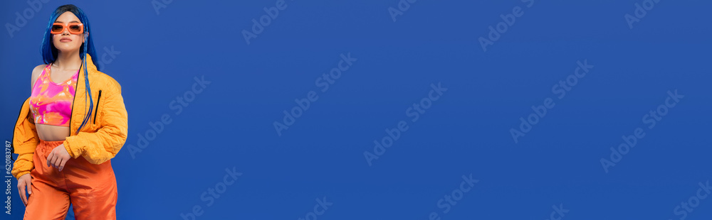 gen z, fashion statement, female model with blue hair adjusting trendy sunglasses isolated on blue background, rebel style, colorful clothes, individualism, young woman, banner