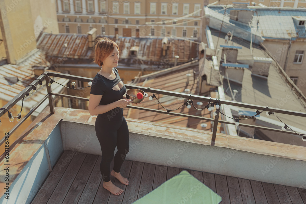 Young woman in black sportswear with Tibetan singing bowl and hammer stands on terrace of roof of city and meditates, concept yoga and meditation in urbanistic city