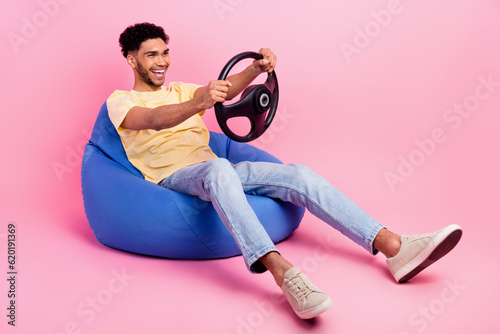Full body photo of young crazy guy driving his rent automobile simulator app hold steering wheel sit pouf isolated on pink color background