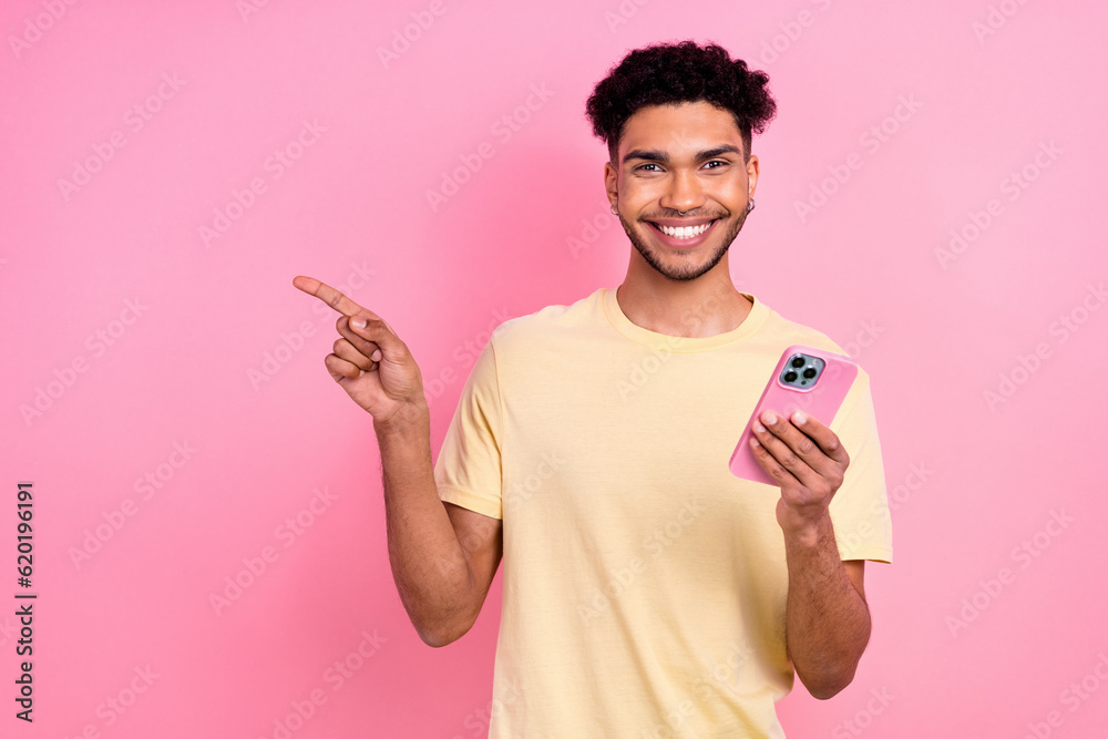 Photo of funny guy afro coiffure dressed yellow t-shirt hold smartphone indicating at eshop empty space isolated on pink color background Photo of funny guy afro coiffure dressed yellow t-shirt hold smartphone indicating at eshop empty space isolated on pink color background