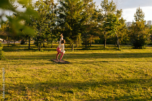 Remote side view of unrecognizable sporty young woman in sportwear doing Extended Side Angle pose during yoga class on evening in nature. Concept of outdoors yoga, fitness, active and sporty lifestyle photo