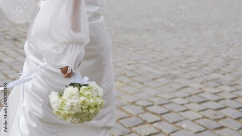 A woman in an elegant, white, wedding, beautiful dress walks along the paving stones with a bouquet of white roses in her hand through the old city. Happy walking down the summer street after a date. 