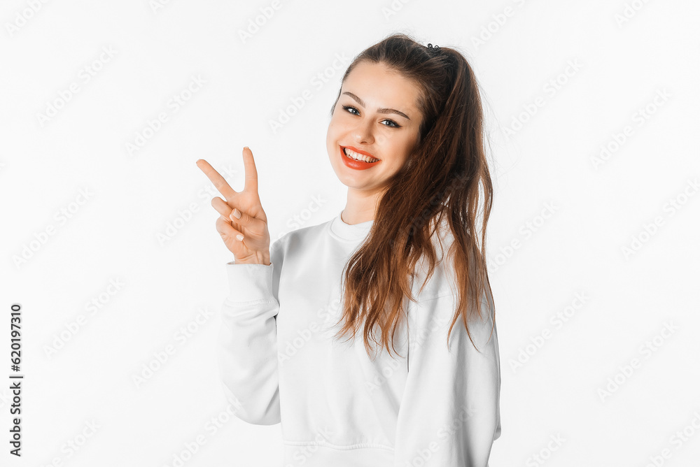 Positive charming brunette girl shows peace sign, makes victory gesture, having fun, posing against white studio background Positive charming brunette girl shows peace sign, makes victory gesture, having fun, posing against white studio background