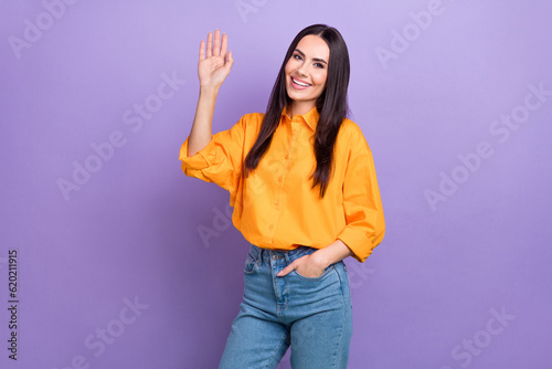 Photo of positive cheerful friendly business woman wear orange shirt denim jeans waves palm greetings isolated on violet color background