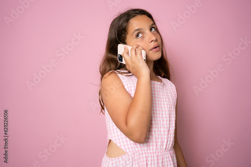 Cute little girl calling on cell phone, good cellular, comfortable to use children mobile device, wearing pink dress. Indoor studio shot isolated on pink background. photo