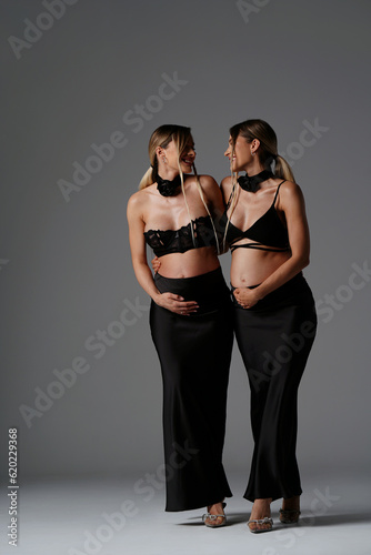 Happy, beautiful pregnant twins sisters in black fashionable and elegant clothes posing together, smiling and touching their bellies. Young women expecting babies.