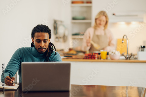 An Arabic freelancer is sitting at home and working on a project on a laptop at the dining table while his spouse is cooking dinner.