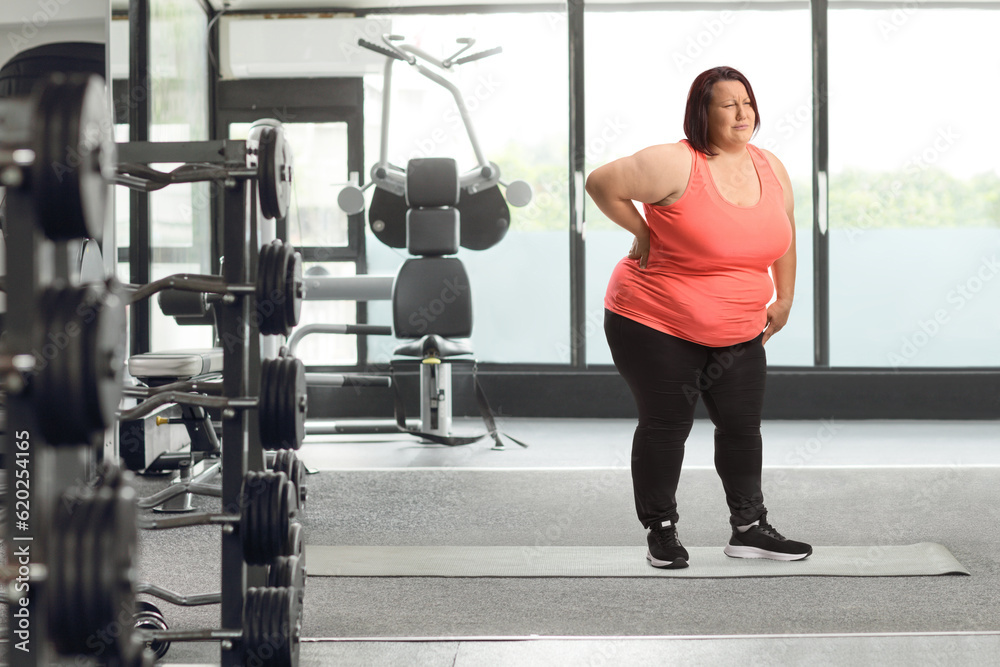 Overweight young woman holding her painful back at the gym Overweight young woman holding her painful back at the gym