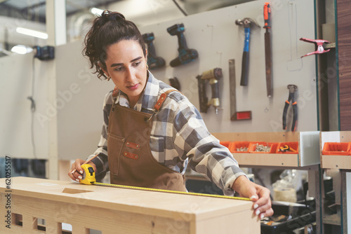 Schilderij op canvas Young woman carpenter using measuring tape at carpentry workshop