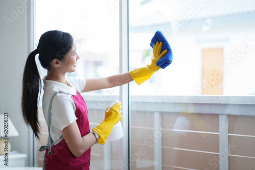 Young housekeeper washing window glass with rag and spray detergent, Positive asian woman doing professional house cleaning, providing sanitary service.