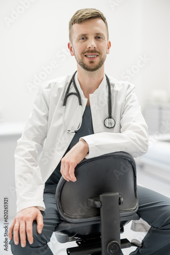 Portrait of young male doctor or practitioner in medical gown with stethoscope sitting on chair at office