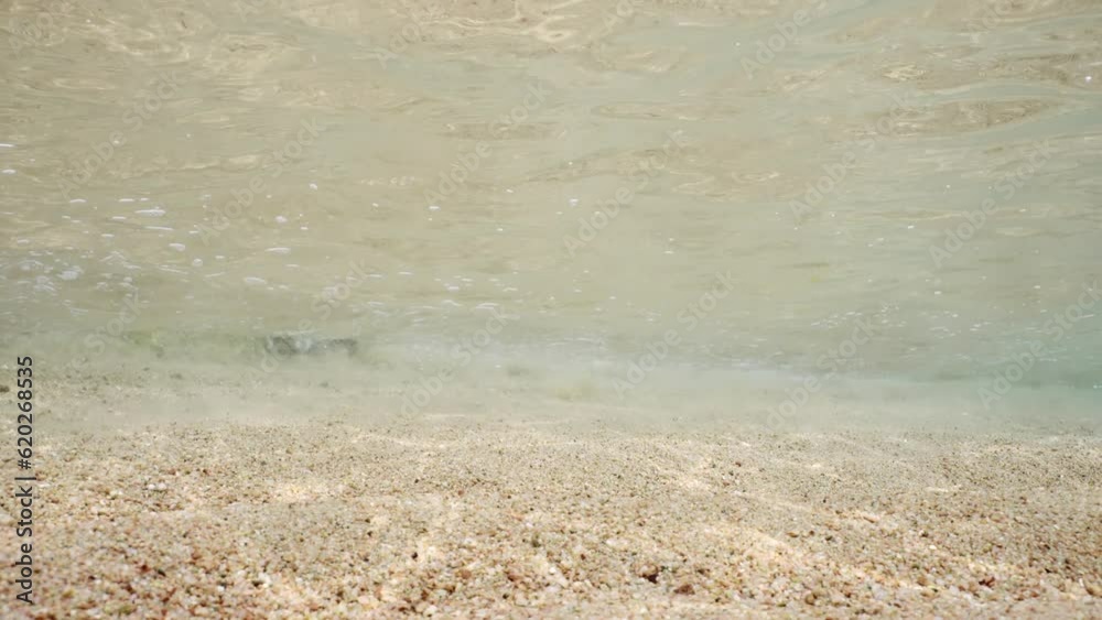 Underwater waves near sandy shore on bright sunny day in sunbeams. Sea surf in coastal area in sandy bottom. Underwater vew of little wave goes out to sand beach on shallow water, slow motion. 