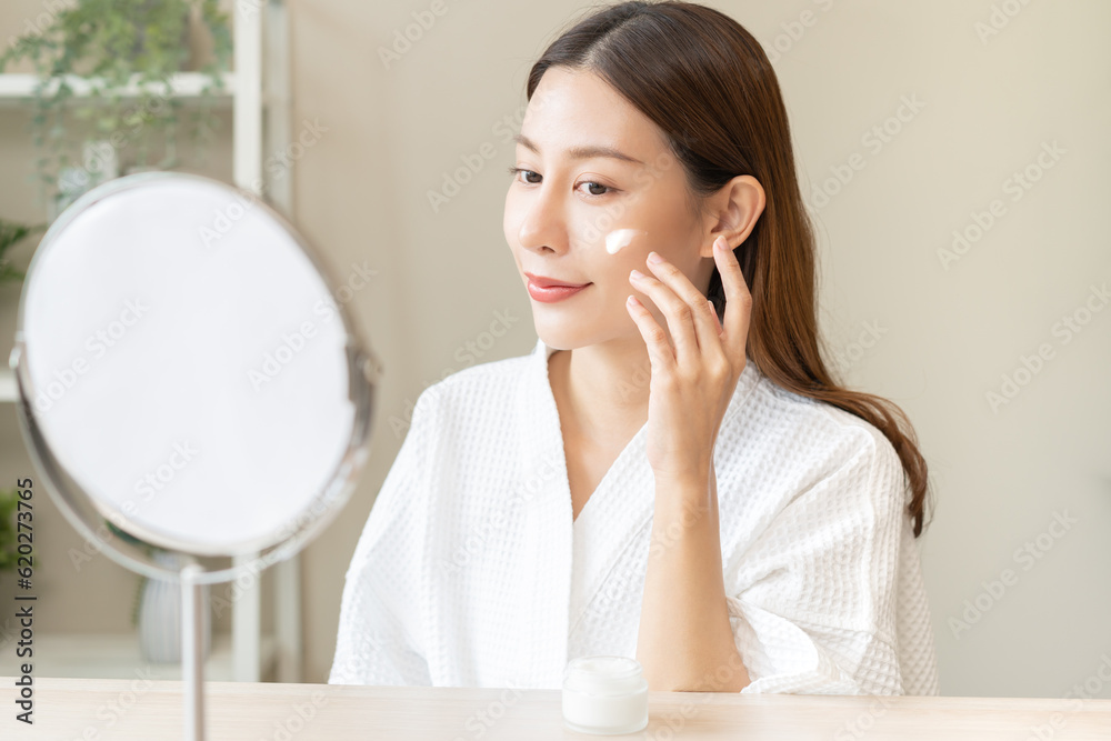 Facial beauty skin care, smile of pretty asian young woman in bathrobe looking at mirror, hand applying moisturizer lotion on her face, putting cream treatment before makeup cosmetic routine at home. Facial beauty skin care, smile of pretty asian young woman in bathrobe looking at mirror, hand applying moisturizer lotion on her face, putting cream treatment before makeup cosmetic routine at home.