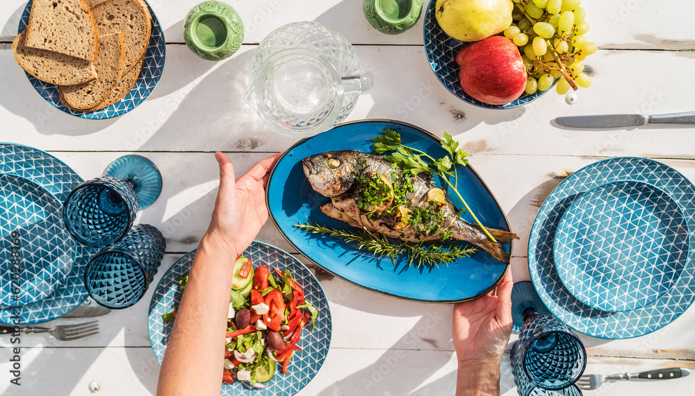Obraz premium Gilt-head bream also known as Orata grilled on a barbeque grill served by a woman on a white wooden table with vegetable salad and fruits. Healthy seafood preparation concept photo.