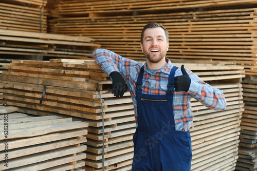 Portrait of a handsome worker choosing the best wooden boards. Carpenter standing next to a big stack of wood bars in a warehouse.