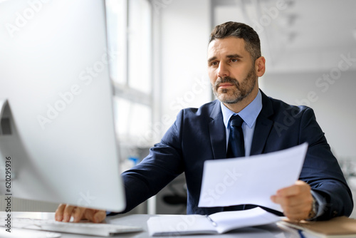 Busy middle aged male entrepreneur working on computer with documents, checking company financial reports