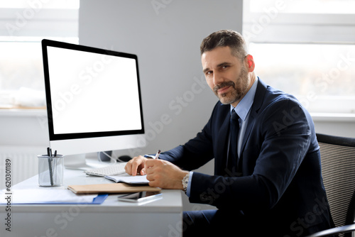 Cheerful middle aged businessman sitting at workplace with computer with blank screen, free space, mockup template
