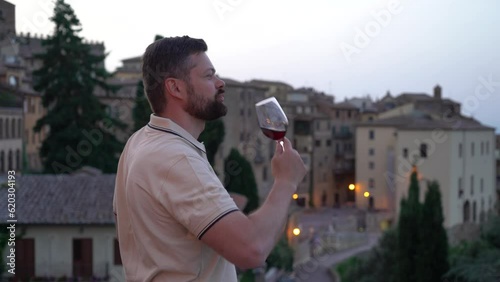 Montepulciano, Province of Siena, Italy. Handsome bearded man drinking red wine and looking at ancient italian city at the evening