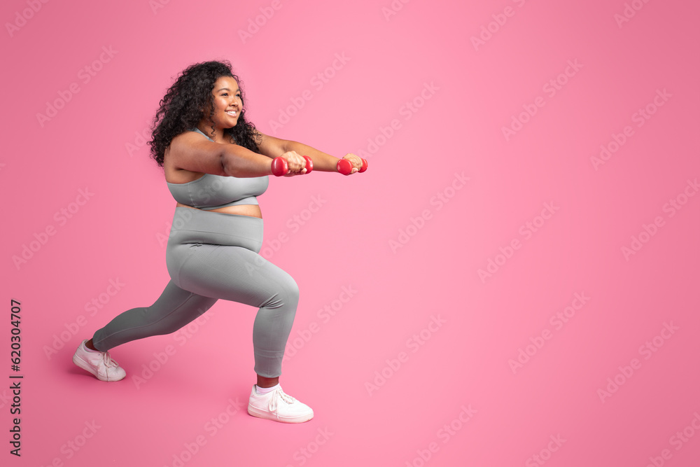 Black chubby woman holding dumbbells and making lunges exercise, training legs over pink studio background, copy space