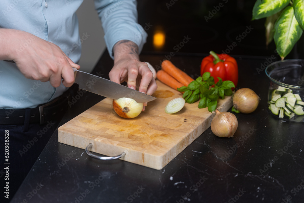 Man in blue shirt cutting and slicing onion og wooden cutting board, with other vegetables in the background. Man in blue shirt cutting and slicing onion og wooden cutting board, with other vegetables in the background.