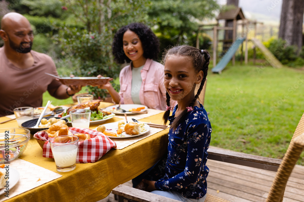 Portrait of happy biracial daughter at table having meal with parents in garden